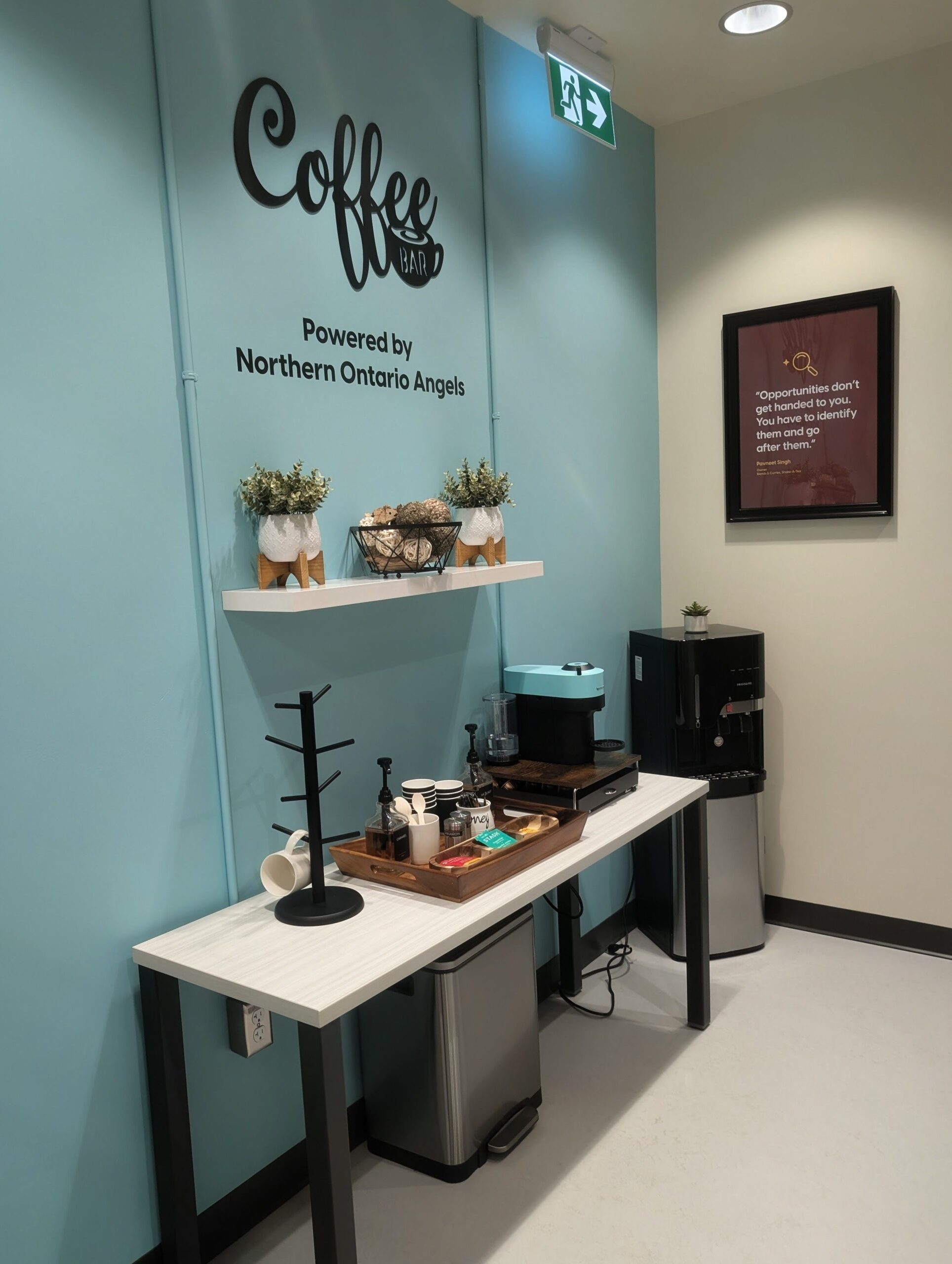 A small coffee station at Hanson Venture Lab features a coffee maker, paper cups, supplies, and a water cooler against a light blue wall with "Coffee Bar" signage and a framed quote.