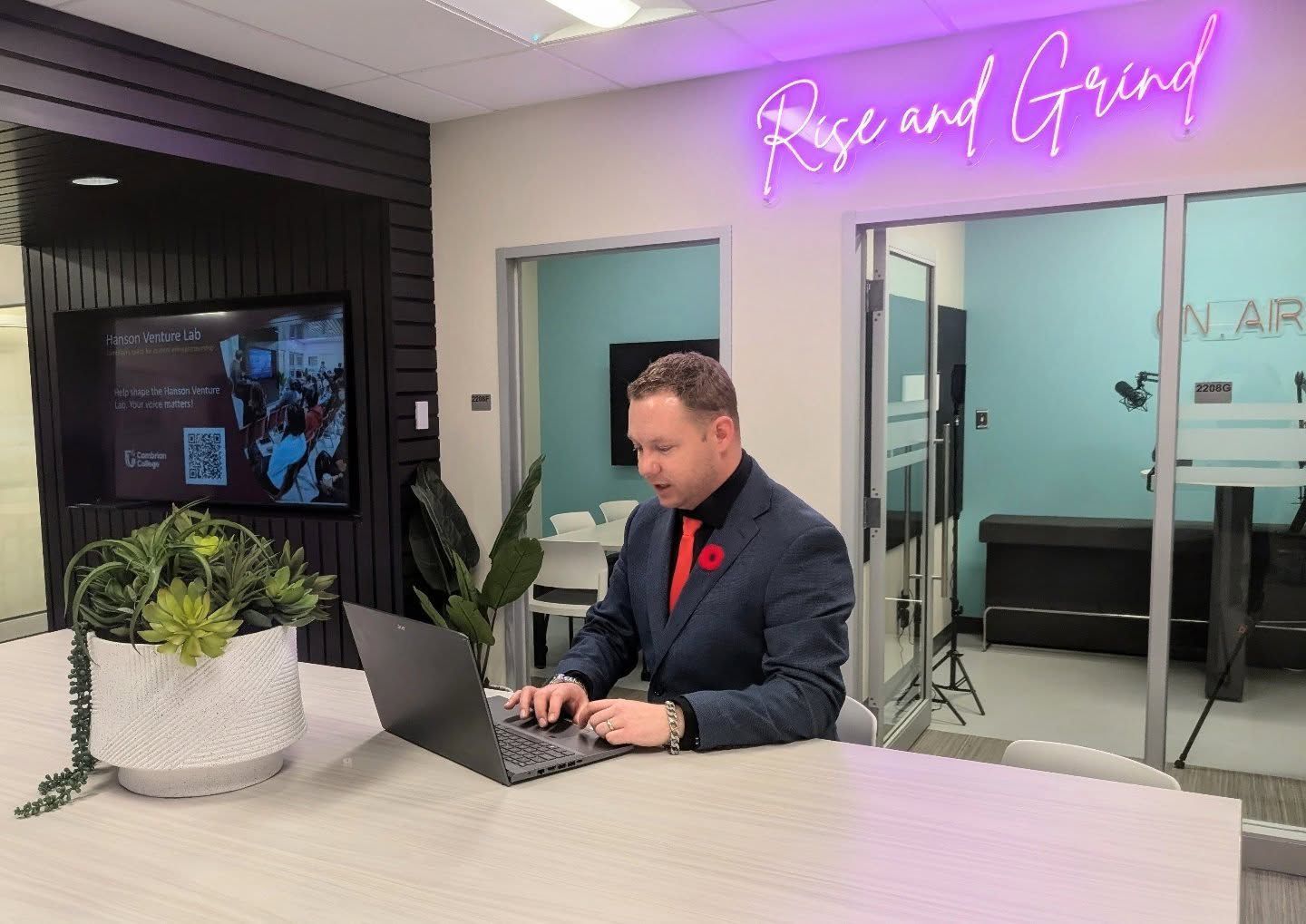 A person in a suit works on a laptop at a modern Hanson Venture Lab office table with plants. A neon sign above reads "Rise and Grind," while a digital display is visible on the wall in the background.