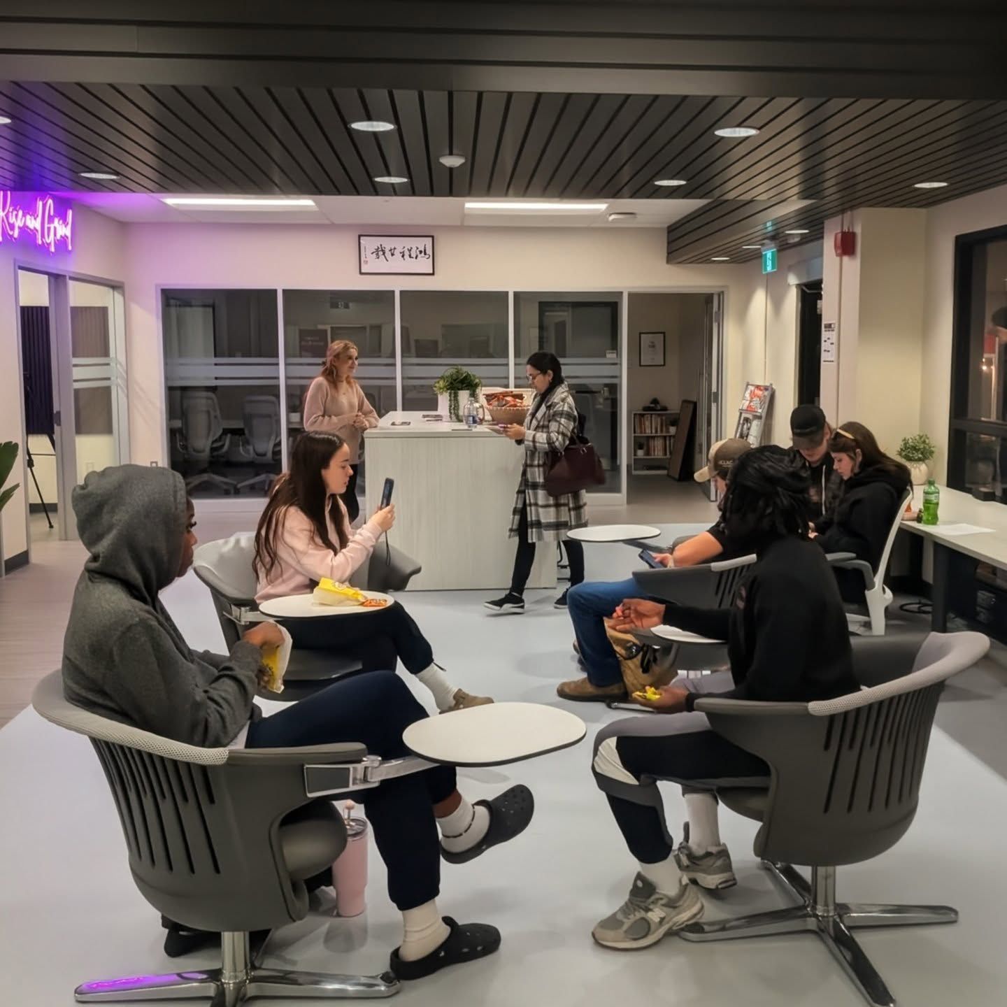 Several people sit in the Hanson Venture Lab’s waiting area with modern chairs, some eating or using phones, while two people interact at a reception desk in the background.