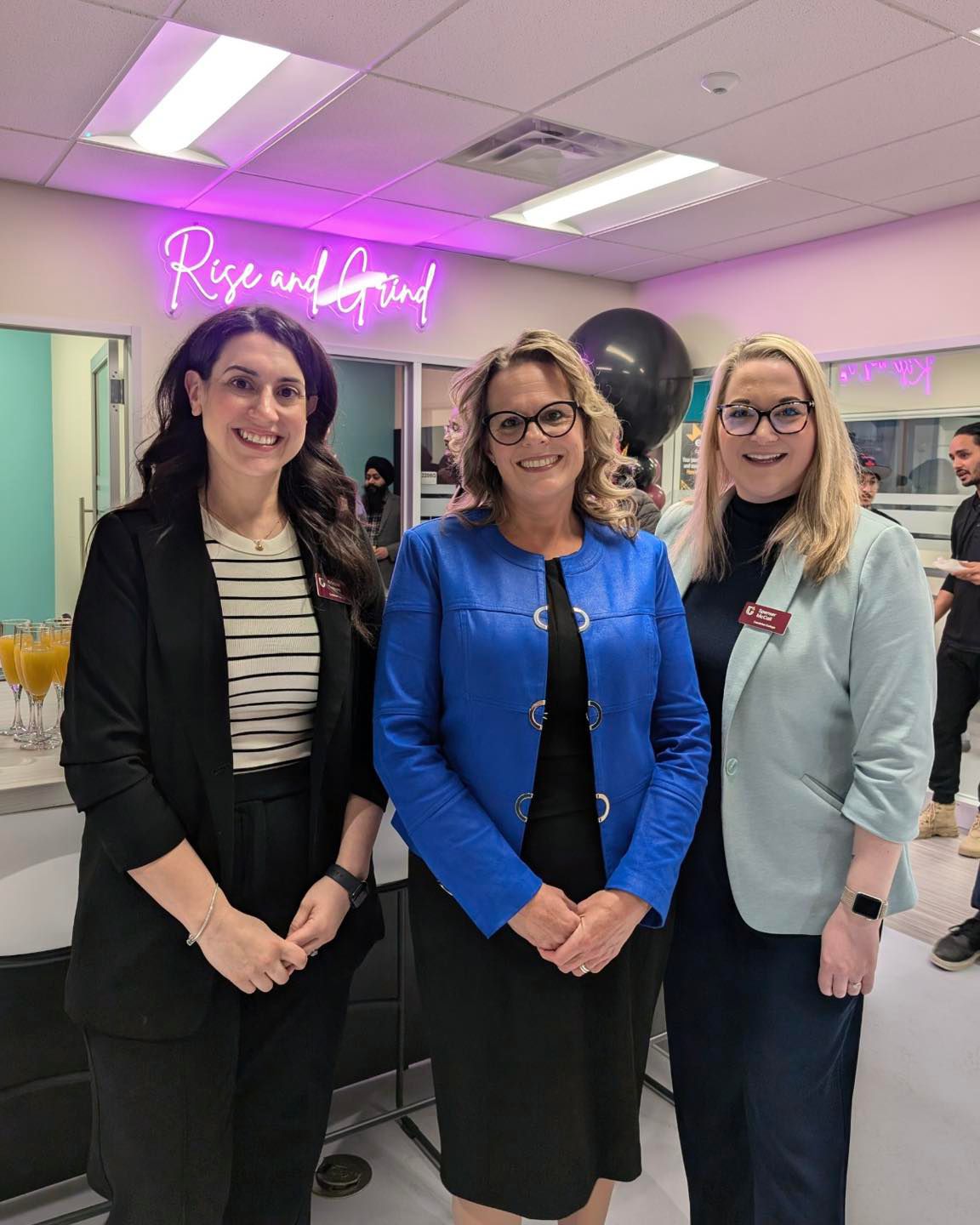 Three people in business attire stand together in a brightly lit room at Hanson Venture Lab, with a "Rise and Grind" neon sign glowing in the background.