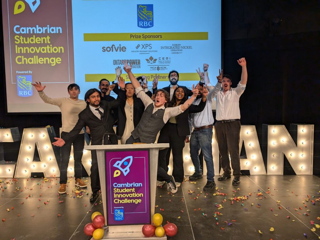 A group of people celebrate on stage at the Cambrian Student Innovation Challenge, with confetti and balloons on the floor, sponsor logos behind them, and support from Hanson Venture Lab energizing the event.