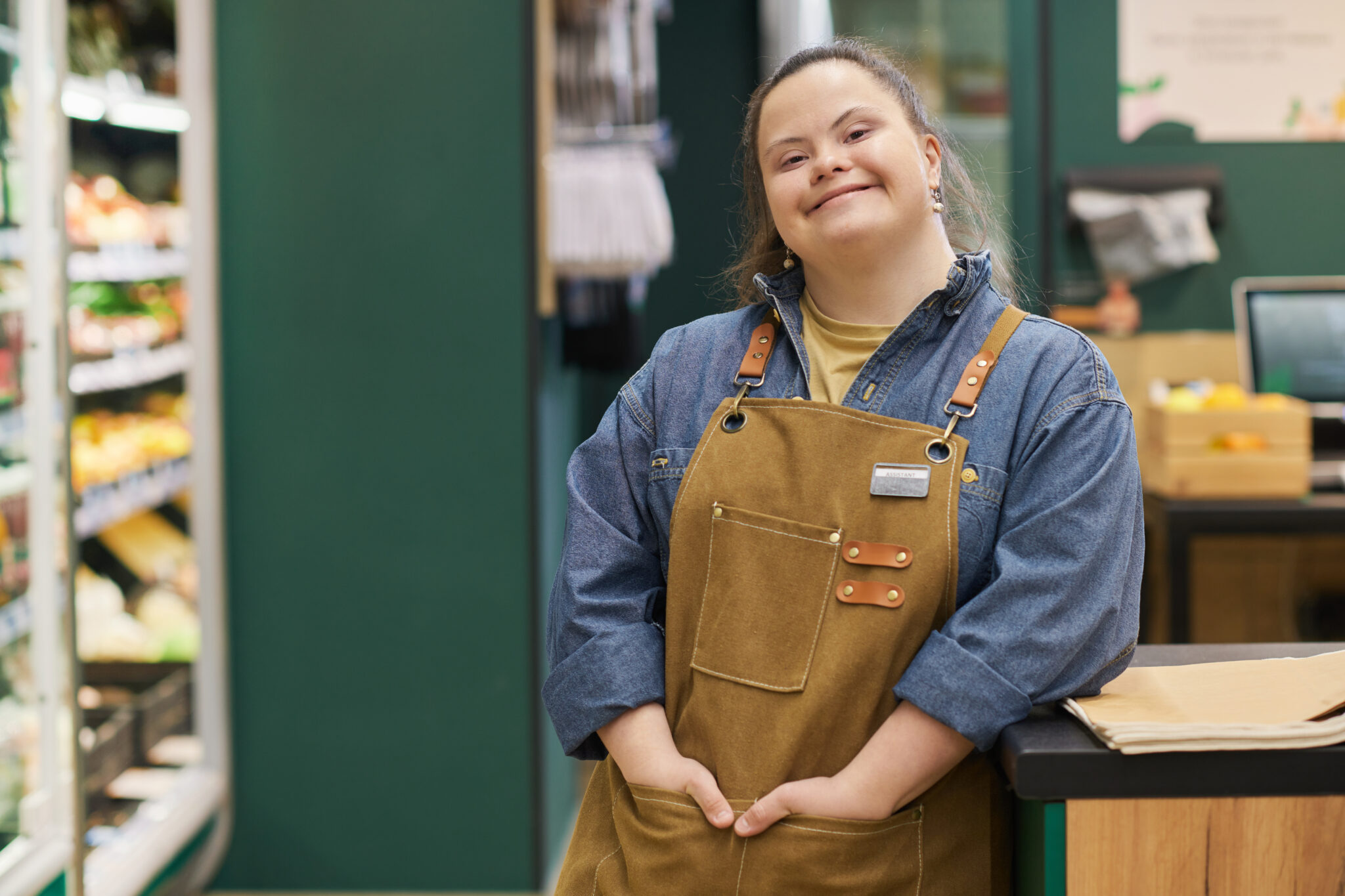 A grocery store employee wearing a brown apron and denim shirt stands by the counter, smiling at the camera—ready to help you build your future with every visit.