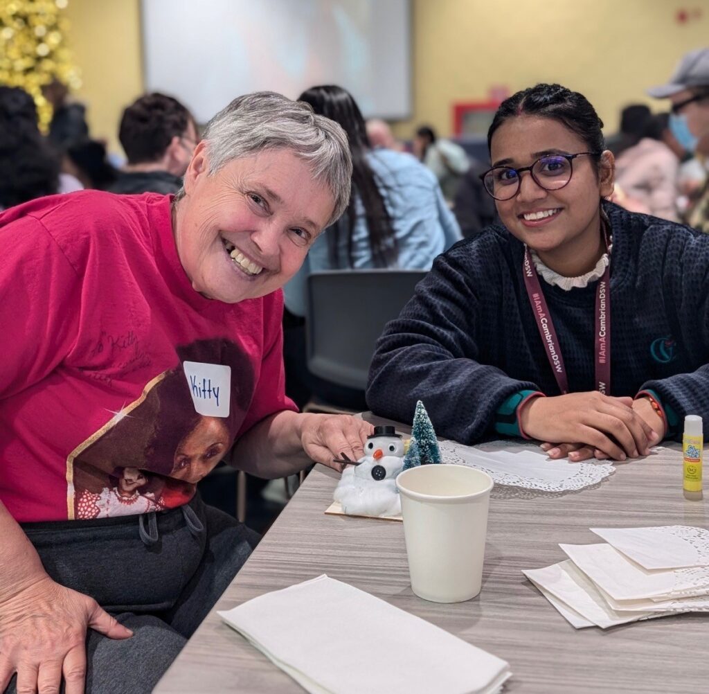 Two people sit at a table with craft supplies, paper napkins, and a cup, smiling at the camera during an indoor activity event.