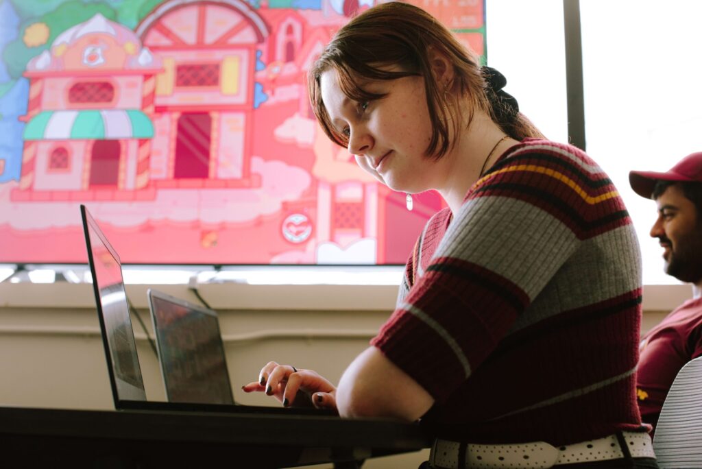 A person with brown hair and a striped sweater works on a laptop at a desk, with a colorful cartoon building displayed on a screen in the background.