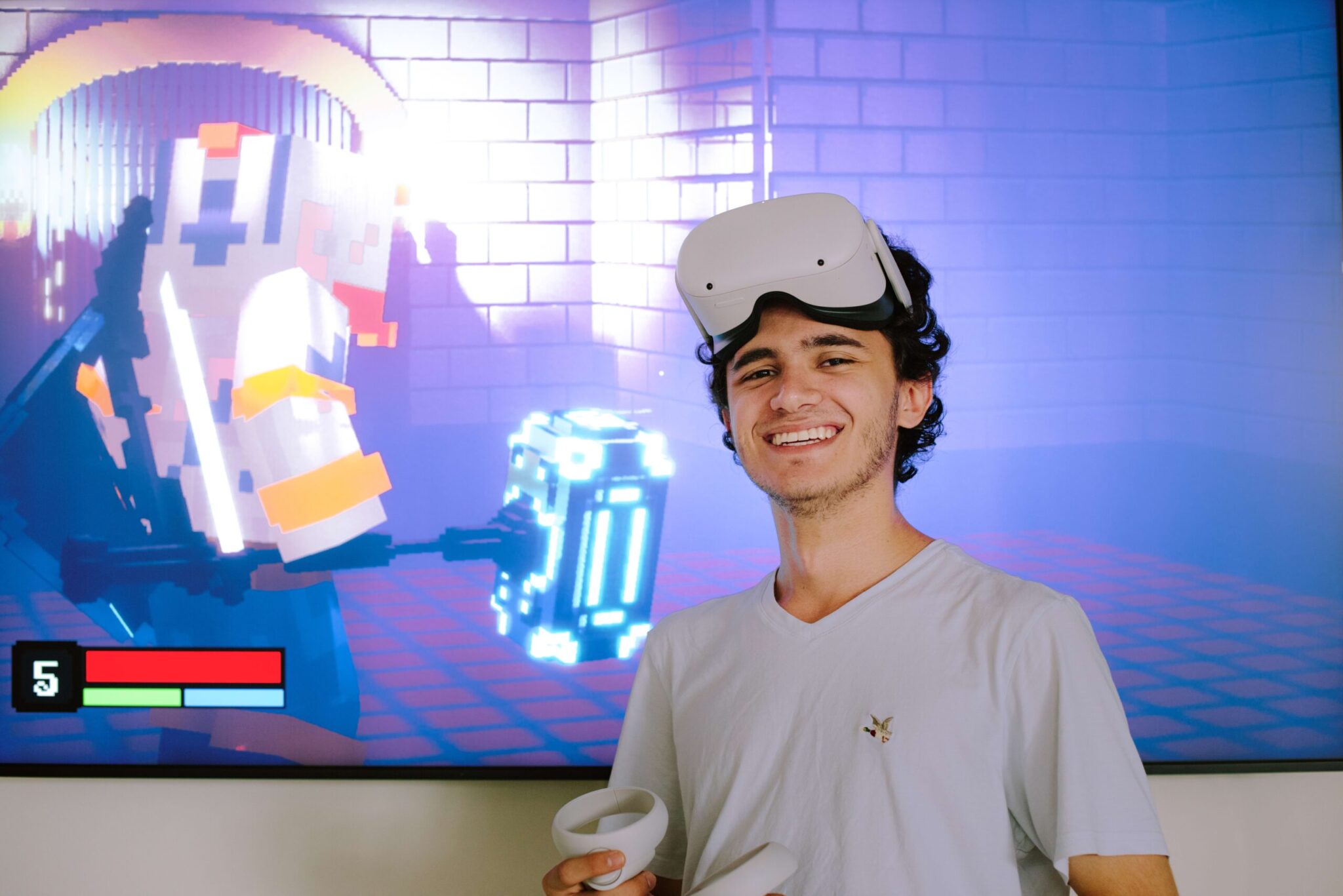 A young man wearing a VR headset and holding controllers stands in front of a screen displaying a pixelated video game character.