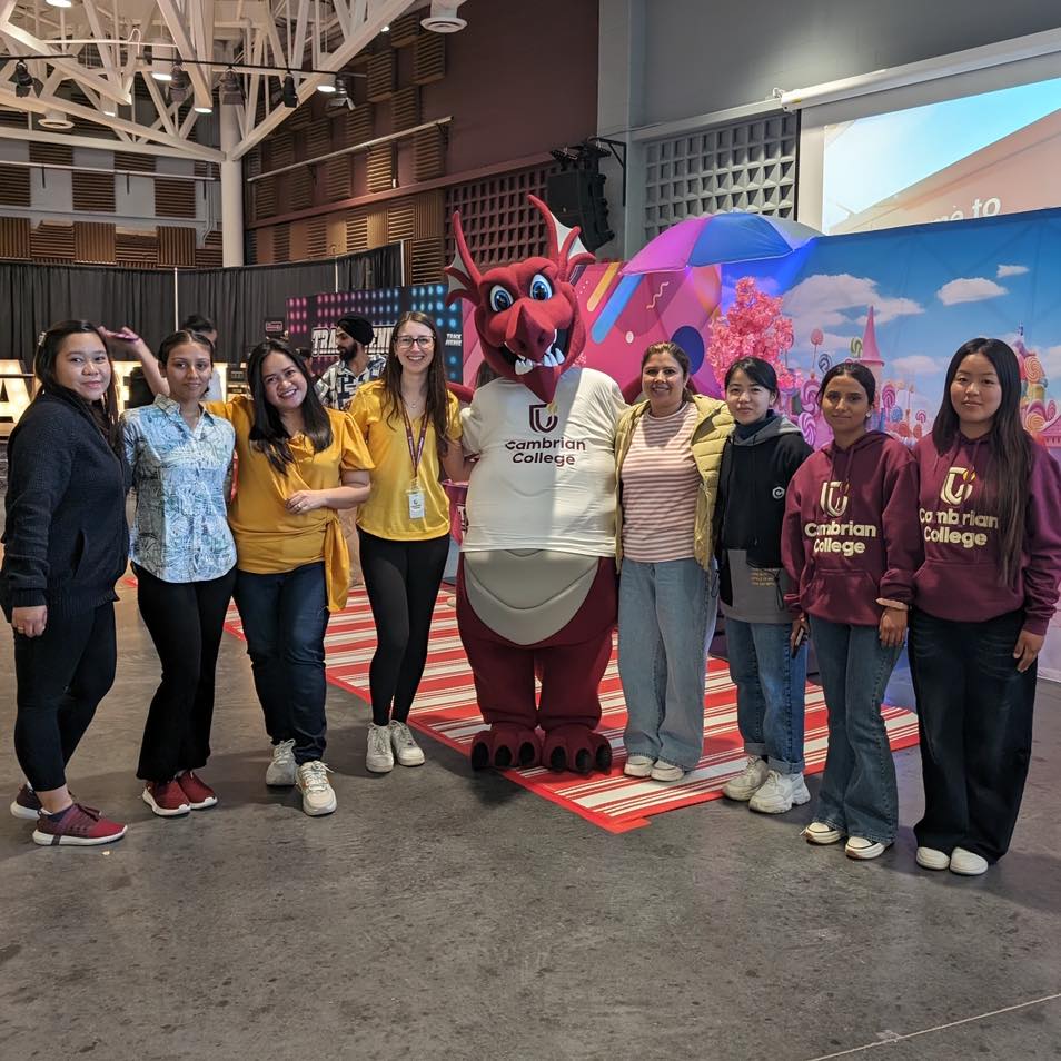 A group of eight people stand indoors next to Burnie wearing a "Cambrian College" shirt; a colorful backdrop is behind them.