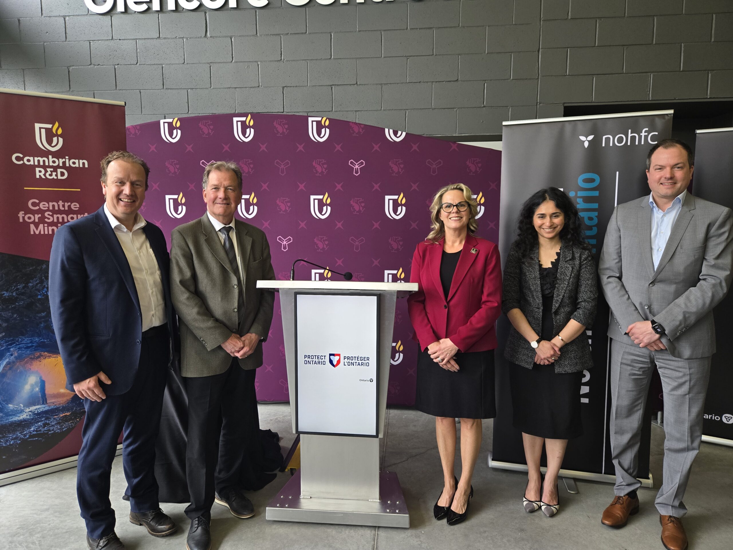 Five people in business attire stand in a row behind a podium, with branded banners from Cambrian College, Ontario, and the Northern Ontario Heritage Fund Corporation highlighting support for critical minerals research in the background.