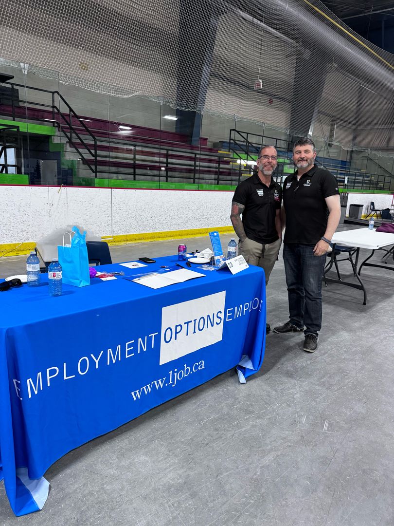 Two men stand behind an Employment Options table at a job fair in an indoor sports arena. The table is covered with informational materials and promotional items.