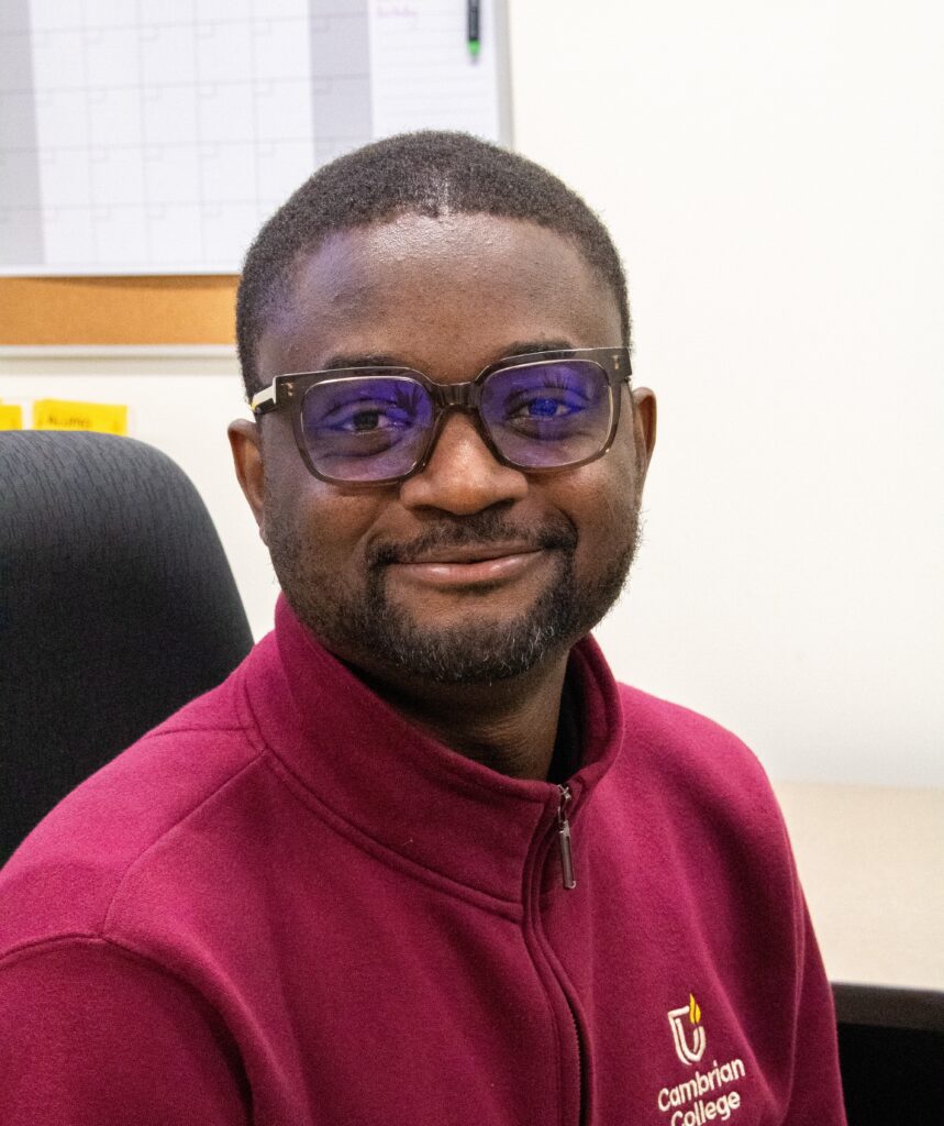 A person wearing glasses and a maroon Cambrian College jacket sits and smiles in an office setting with a calendar on the wall behind him, ready to welcome participants to the International Virtual Open Day Africa.