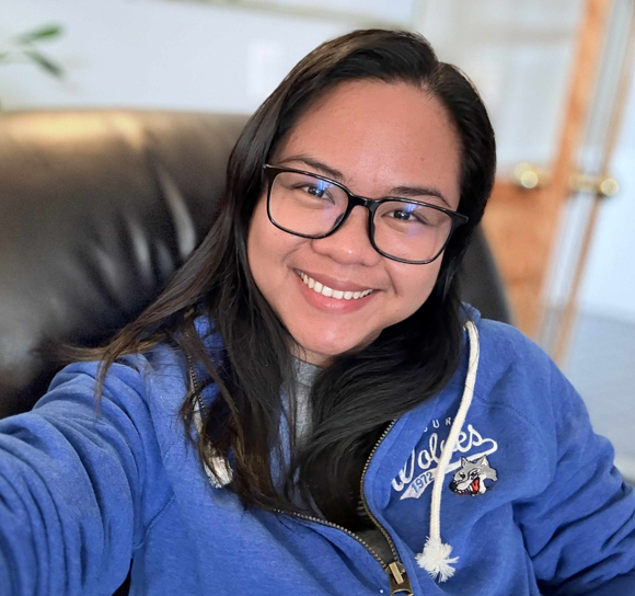 A person with long dark hair and glasses sits on a dark chair, smiling and wearing a blue Wolves hoodie, ready to participate in local Community Programs.