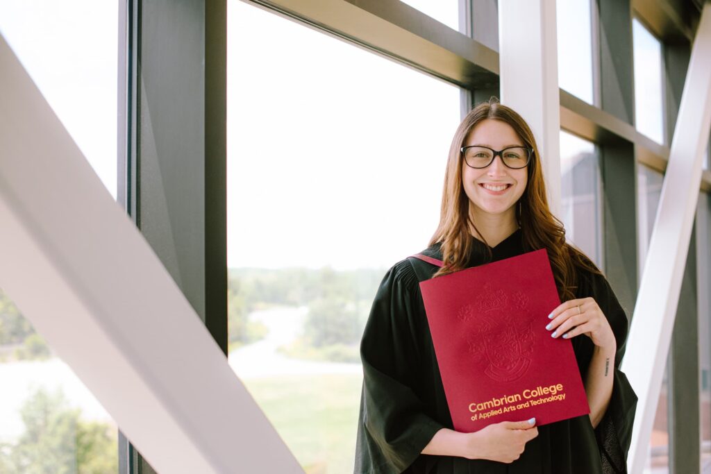 A graduate in a black gown and glasses holds a Cambrian College diploma folder, standing by large windows with trees visible outside.