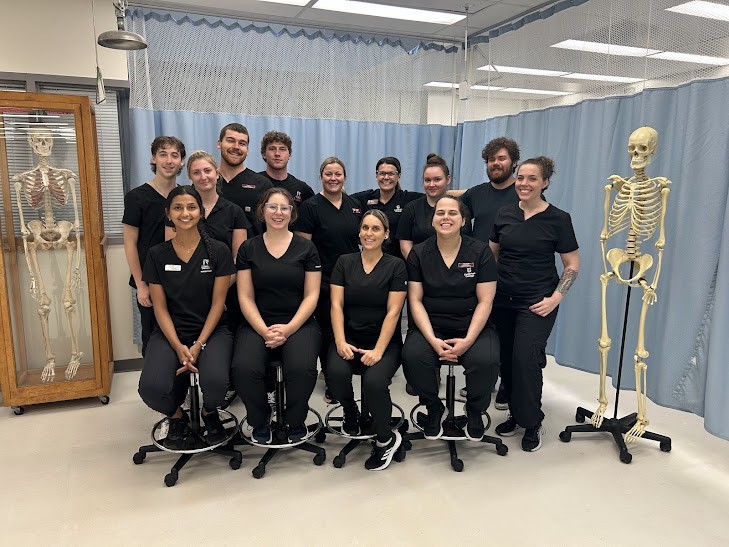 Thirteen people in black scrubs pose for a group photo in a medical or educational setting, flanked by two anatomical skeleton models.