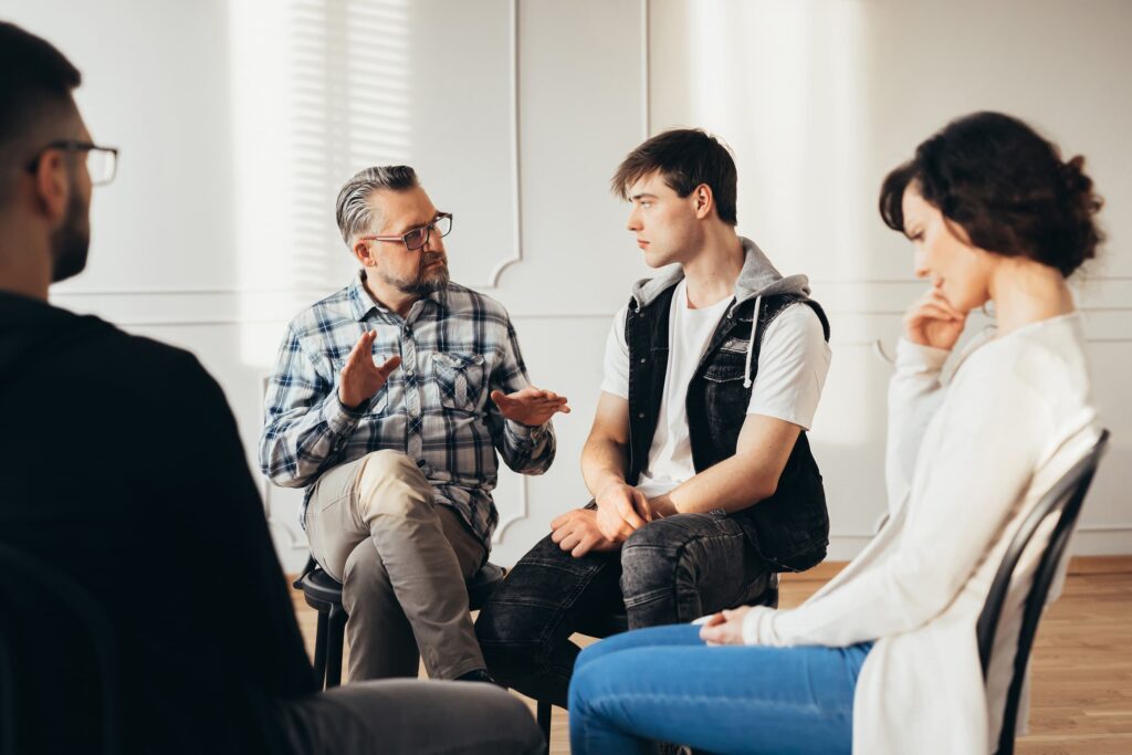 A group of four students sit in a circle having a serious discussion in a brightly lit room. The professor is speaking while the others listen attentively.