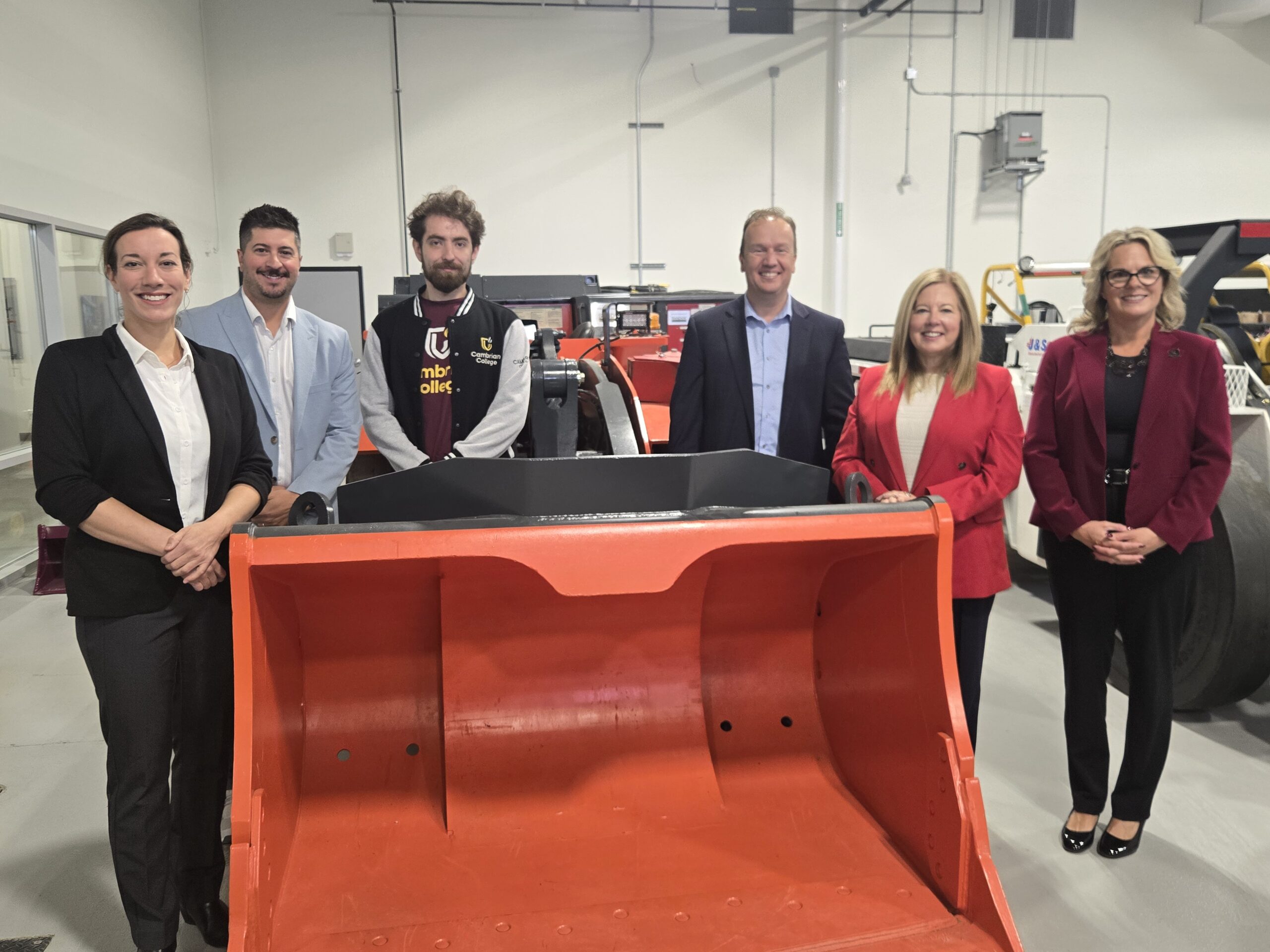 Six people standing indoors behind an orange industrial vehicle, posing for a group photo in a well-lit workshop or facility.