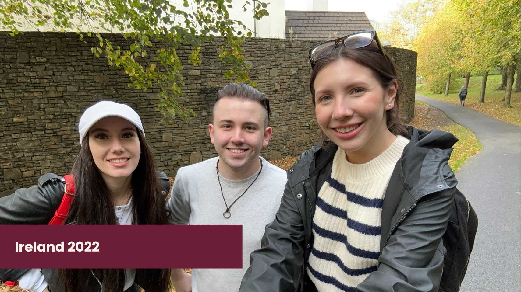 Three students smile at the camera on a paved path lined with autumn trees and a stone wall, with "Ireland 2022" and the Cambrian College Global Skills Opportunity highlighted in the corner.