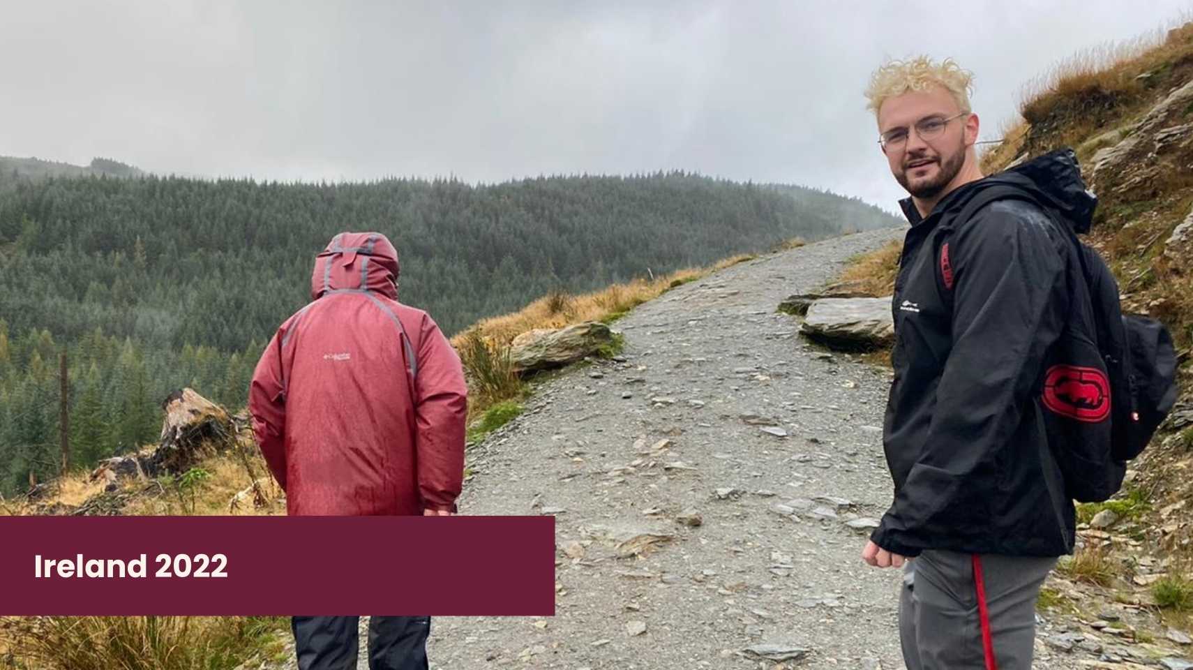 Two students wearing jackets walk on a rocky mountain trail with forested hills in the background. Text overlay reads "Ireland 2022, Cambrian College Global Skills Opportunity.
