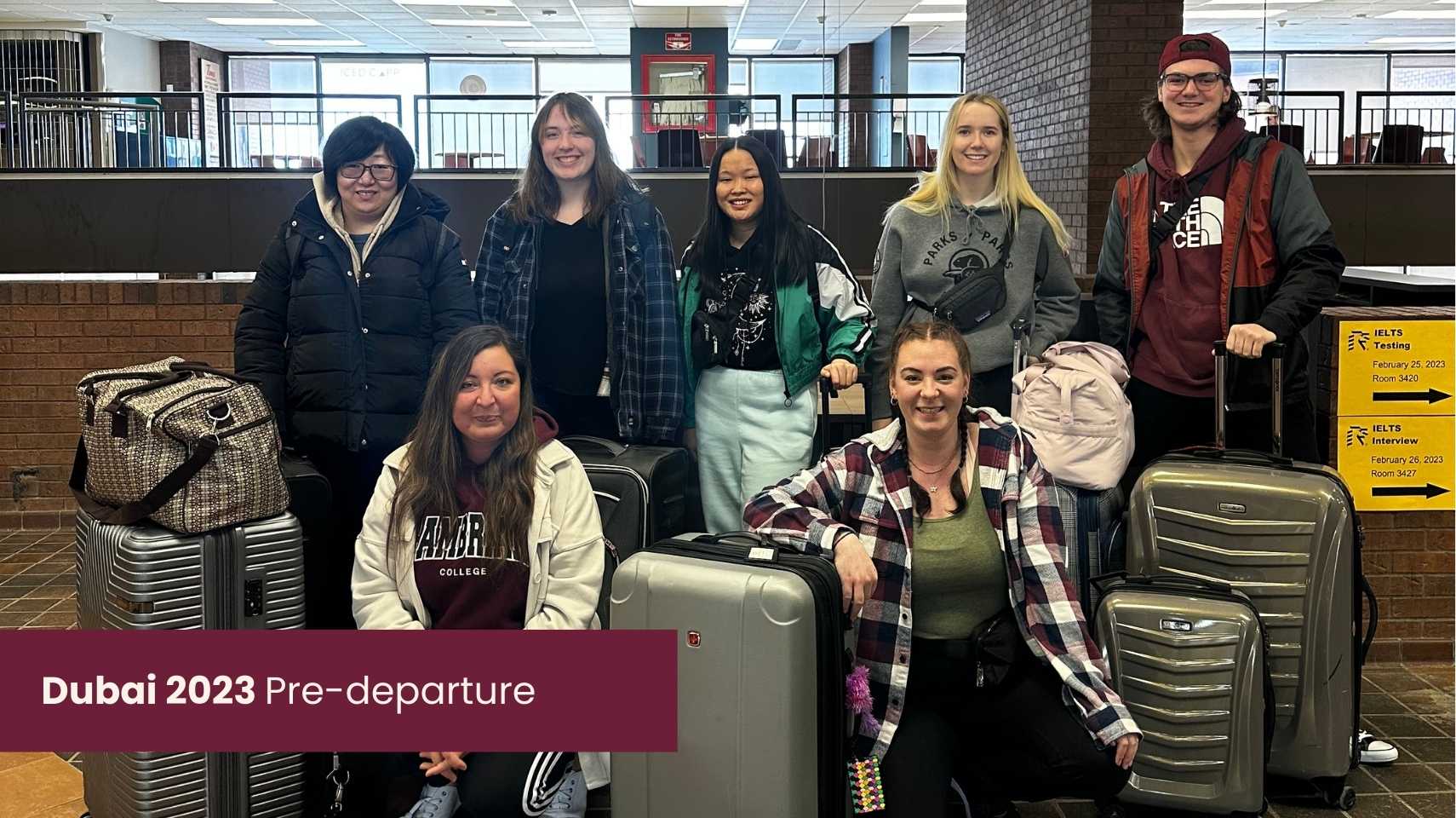 Seven people with luggage pose indoors at what appears to be an airport or transit area, ready for their Cambrian College Global Skills Opportunity. A sign reads "Dubai 2023 Pre-departure.