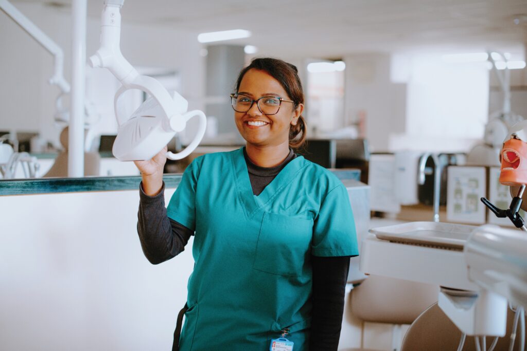 A dental professional in scrubs stands in a dental clinic, smiling and adjusting an overhead examination light.