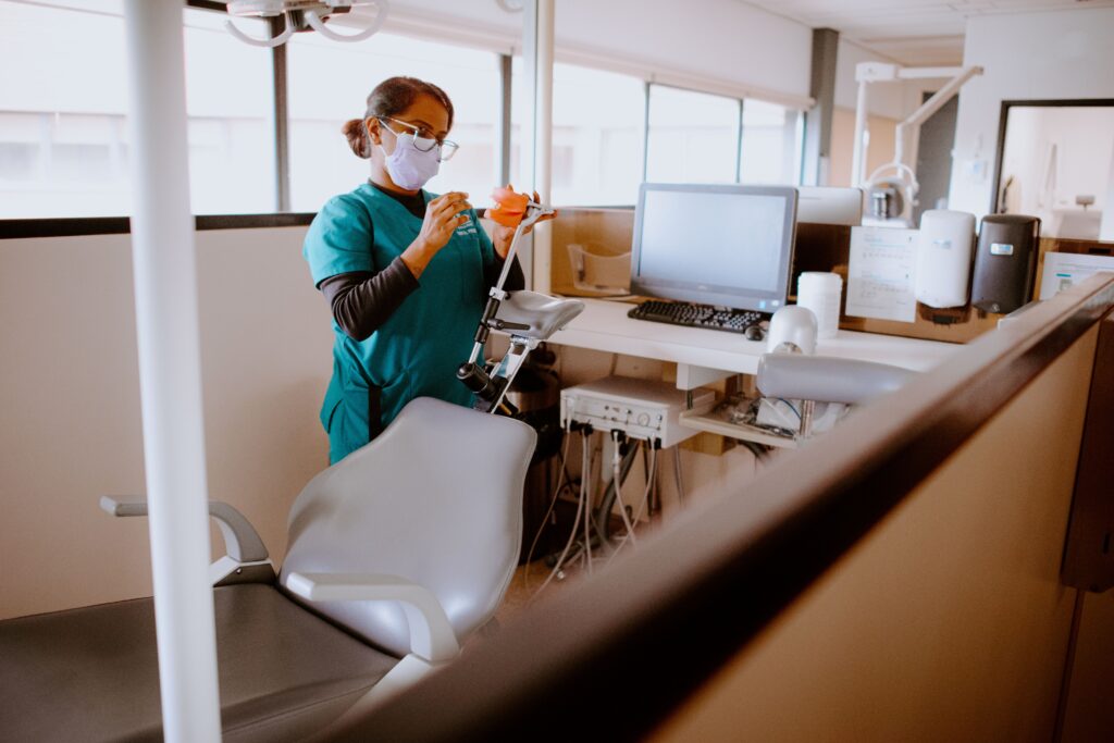 A dental professional wearing scrubs and a mask disinfects a dental chair and equipment in a clinic.
