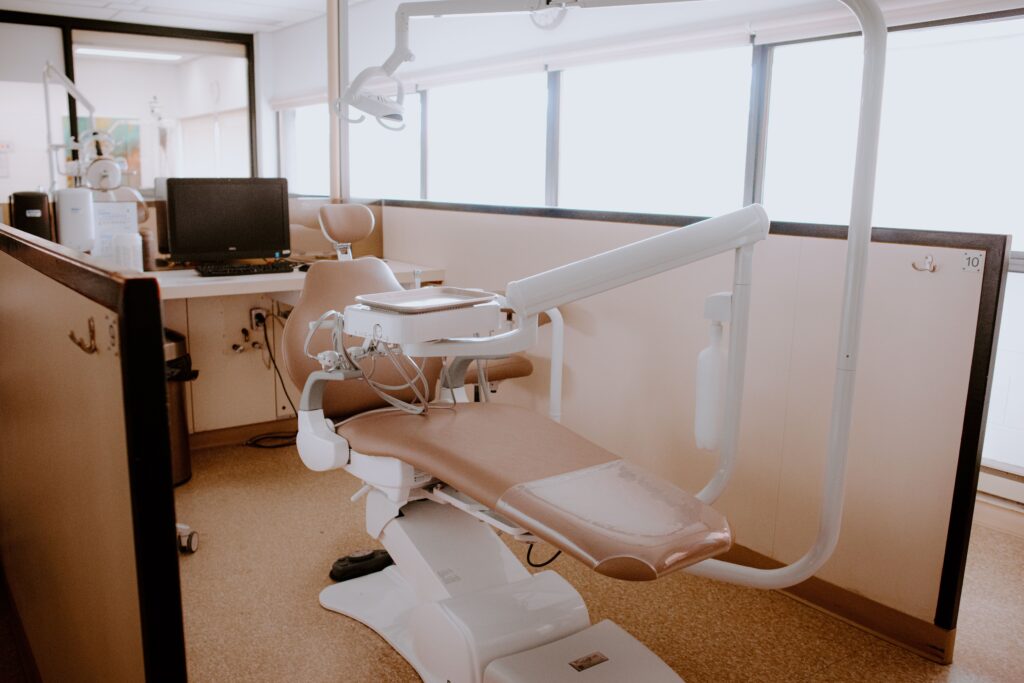 A dental exam room with a reclining dentist chair, overhead light, computer workstation, and beige walls.
