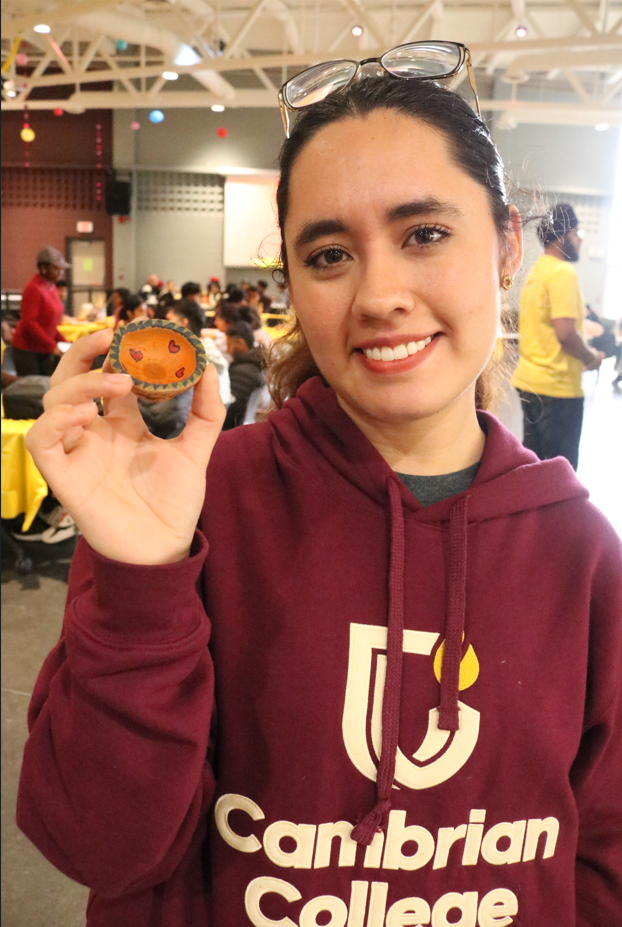 A person in a Cambrian College hoodie holds up a small painted pumpkin with a face on it, standing in a large indoor space with people in the background, getting into the Pre-Party Prep spirit for Diwali.