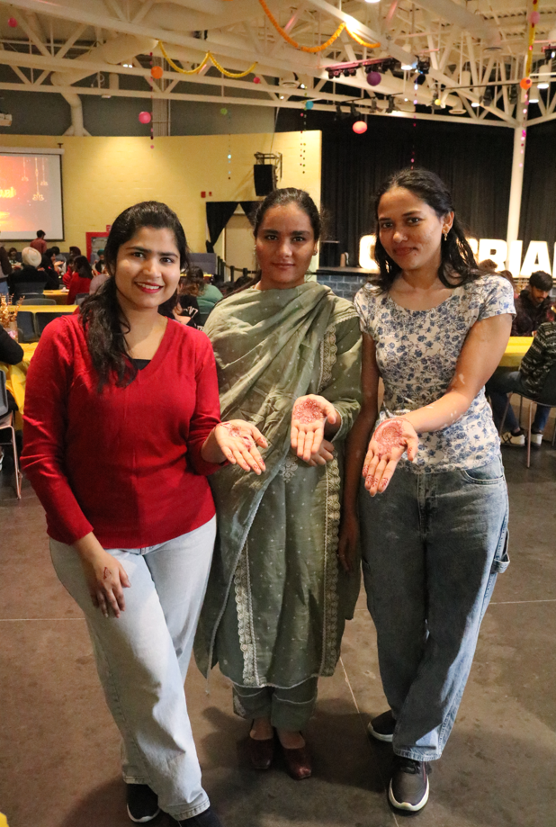 Three people stand indoors, facing the camera and showing intricate henna designs on their palms—a perfect snapshot of Diwali pre-party prep. Tables and festive decorations are visible in the background.
