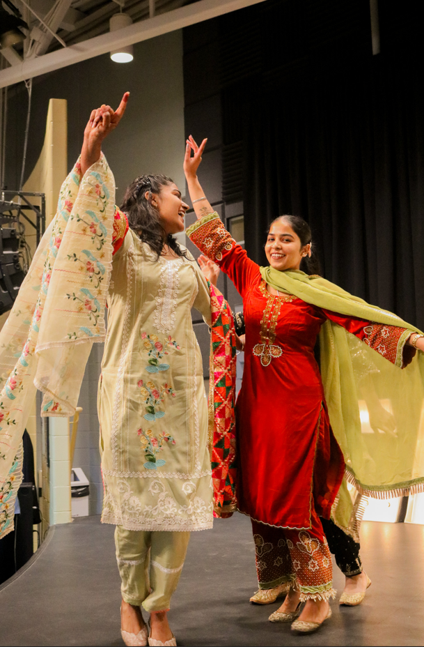Two people in traditional South Asian attire dance joyfully on stage, raising their arms and smiling, celebrating Diwali with vibrant energy. The festive prep scene unfolds against black curtains in the background.