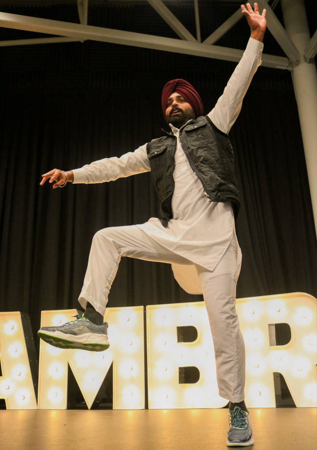 Person in traditional attire and sneakers performs a dance on stage, with one leg raised and arms outstretched, celebrating Diwali in front of illuminated marquee letters.