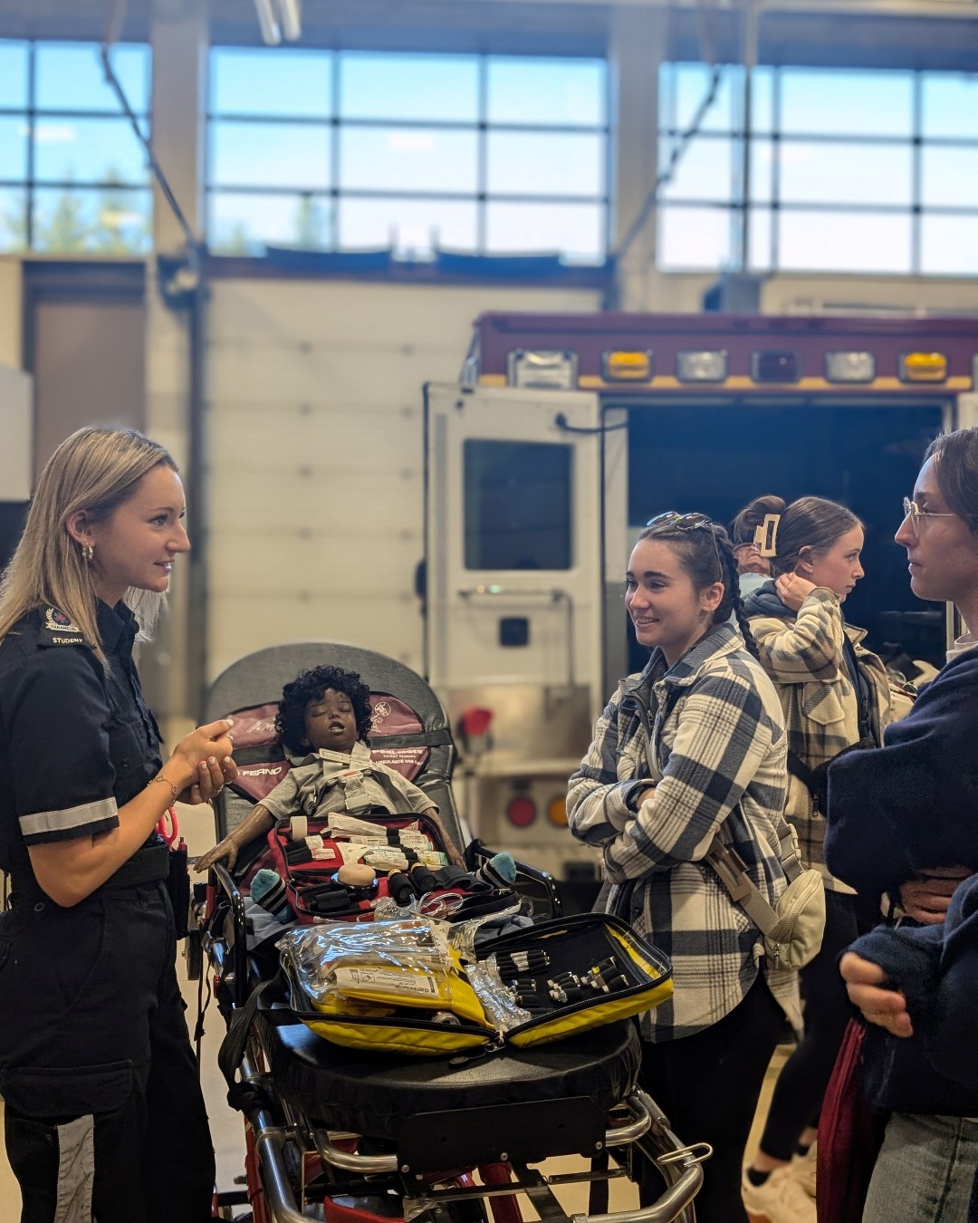 A paramedic explains equipment to three women gathered around a stretcher with a medical mannequin inside a garage bay with an ambulance in the background.