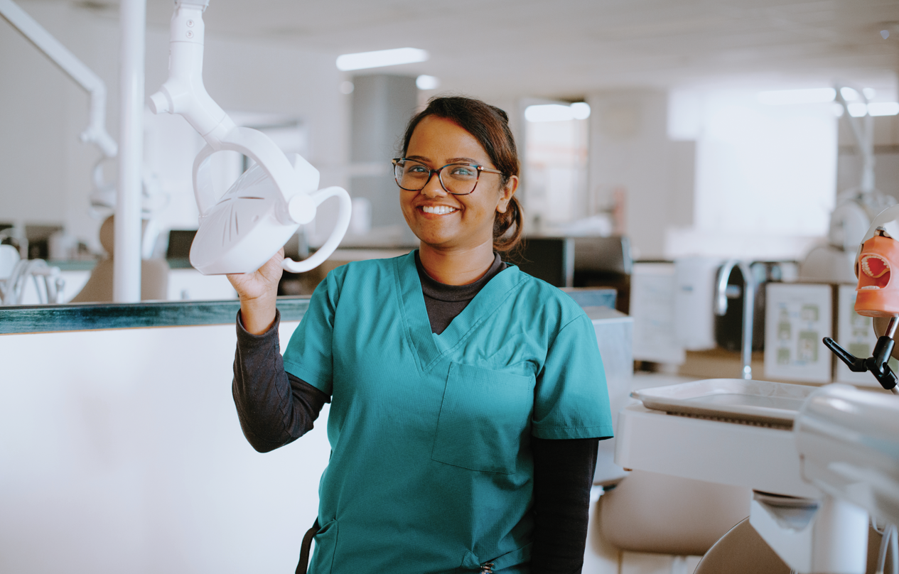 A dental professional in teal scrubs stands in a dental clinic, smiling and holding an overhead dental lamp.