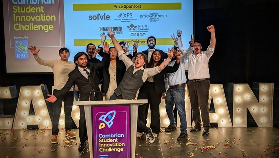 A group of people on stage celebrates at the Cambrian Student Innovation Challenge, standing behind a podium with sponsors’ logos displayed on a screen behind them.