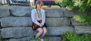 Taylor Fitton, dressed in graduation attire, sits on stone steps outdoors with greenery and parked cars in the background.