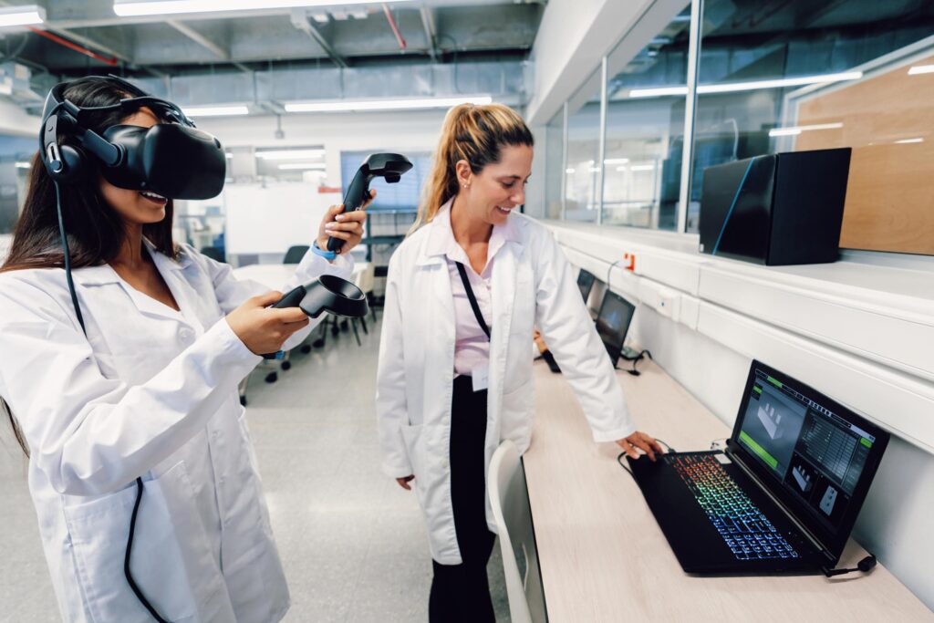 Two students in lab coats use virtual reality equipment in a laboratory; one wears a VR headset and holds controllers, while the other looks at a laptop displaying serious games software for research and development.