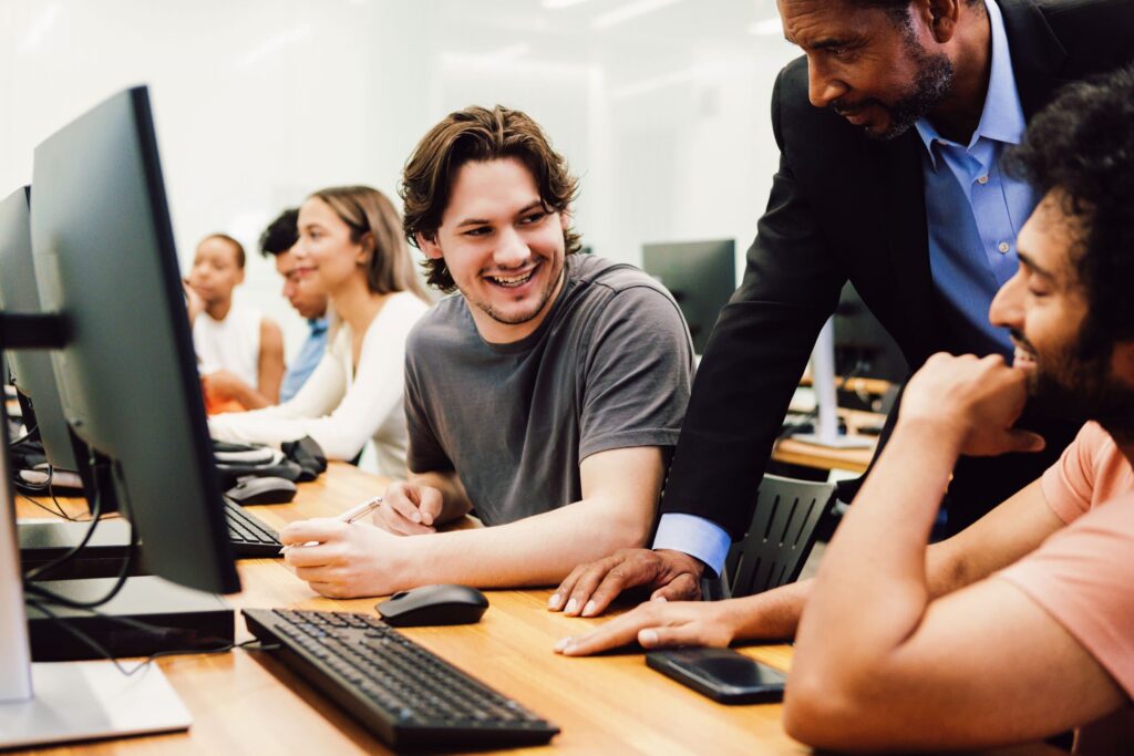 A group of students sit at desks with computers in a classroom; an instructor talks to two Bachelor of Applied Computing students who are smiling and engaged in conversation.