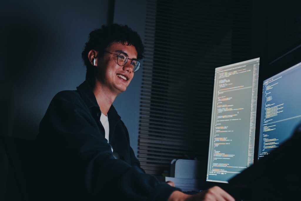 A student wearing glasses and earbuds sits in front of two monitors displaying code in a dimly lit room.