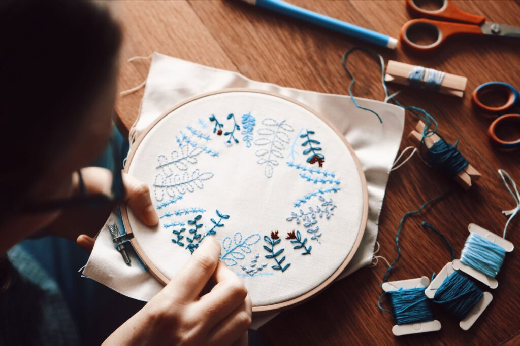 A student is embroidering a floral pattern with blue and green thread on white fabric using an embroidery hoop, surrounded by embroidery supplies on a wooden table.