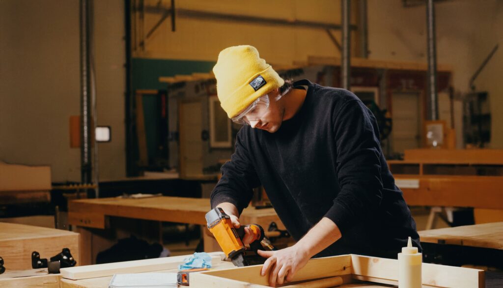 A student wearing a yellow beanie and safety glasses uses a power tool to assemble wooden boards in a trade shop.