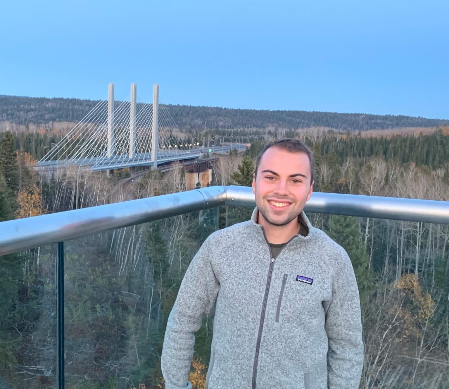 Eric Sampson, a professional in Civil Engineering Technology, stands on a balcony in a gray fleece jacket, overlooking a forested area and a modern cable-stayed bridge at dusk.