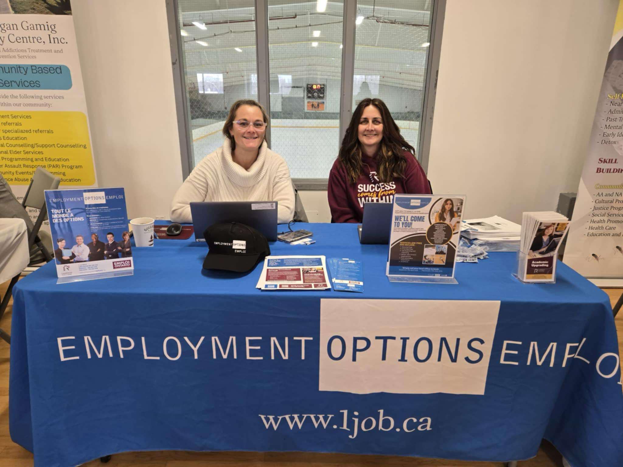 Two people sit at an Employment Options booth with laptops and informational materials on a blue tablecloth, sharing details about Cambrian Satellite Campuses at a community event.