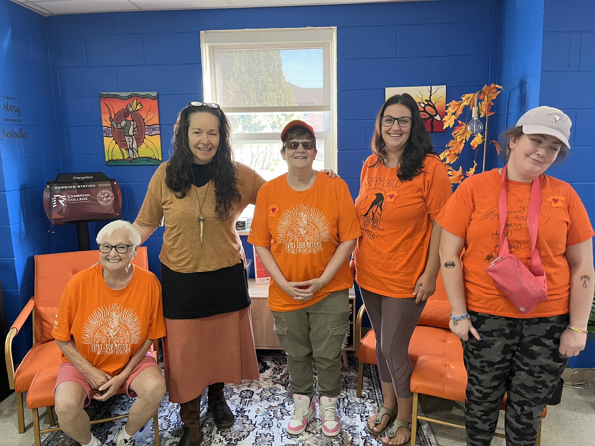 Five women, three in matching orange t-shirts, stand and sit together in a brightly colored room with blue walls and orange chairs at one of the Cambrian Satellite Campuses.
