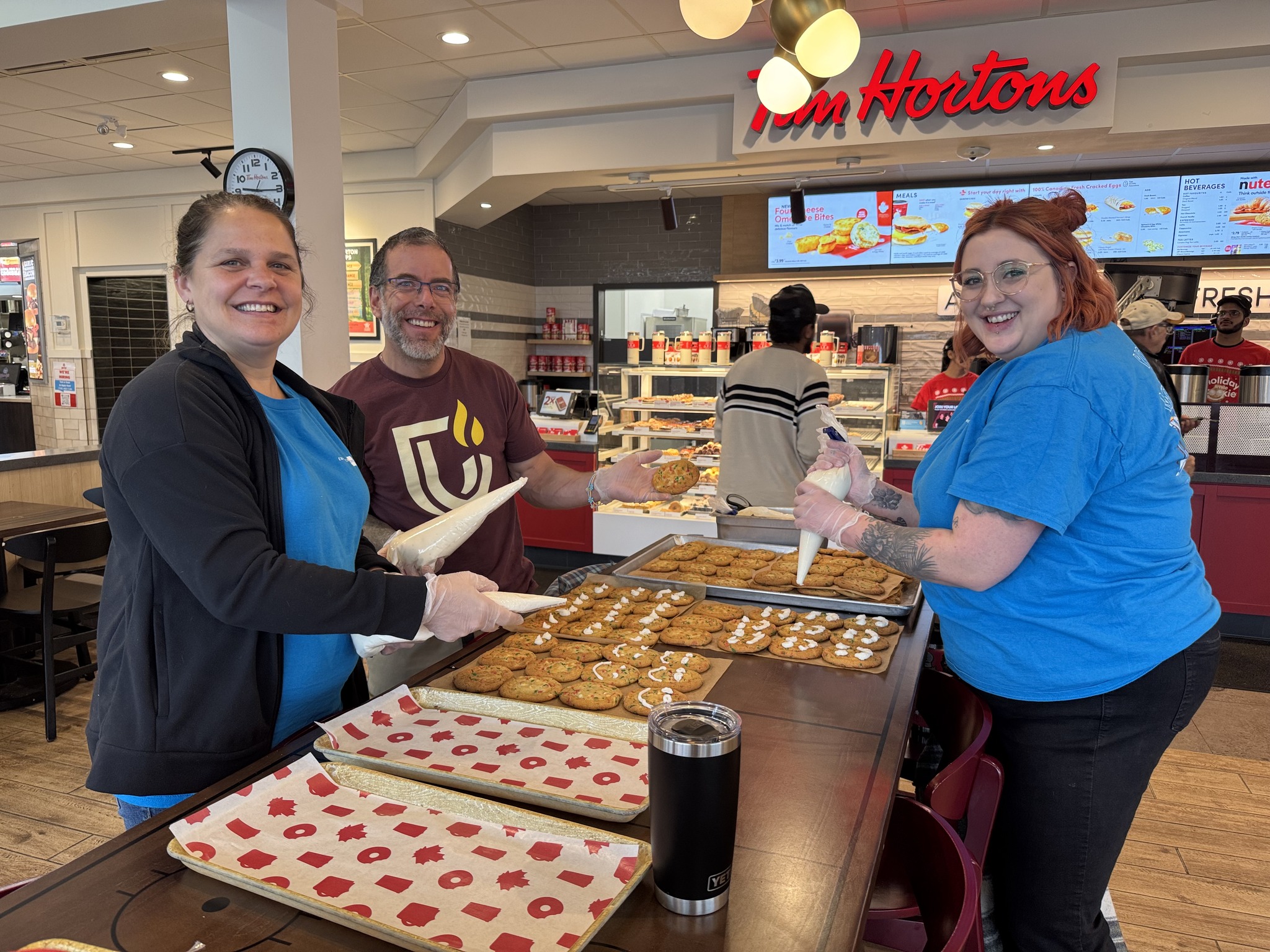 Three people are decorating trays of cookies inside a Tim Hortons cafe at Cambrian Satellite Campuses, smiling at the camera. Baking supplies and finished cookies are on the counter.