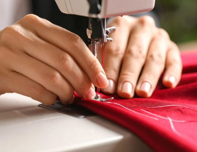 Close-up of hands guiding red fabric through a sewing machine, stitching a seam with white thread. A perfect example of lifestyle learning through Continuing Education.