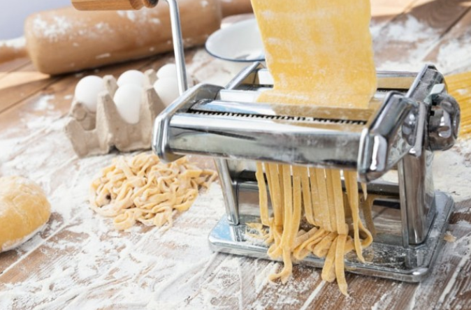 A manual pasta machine rolls and cuts fresh pasta dough on a floured wooden surface, with eggs and a rolling pin nearby. Perfect for anyone exploring leisure learning or continuing education in the culinary arts.