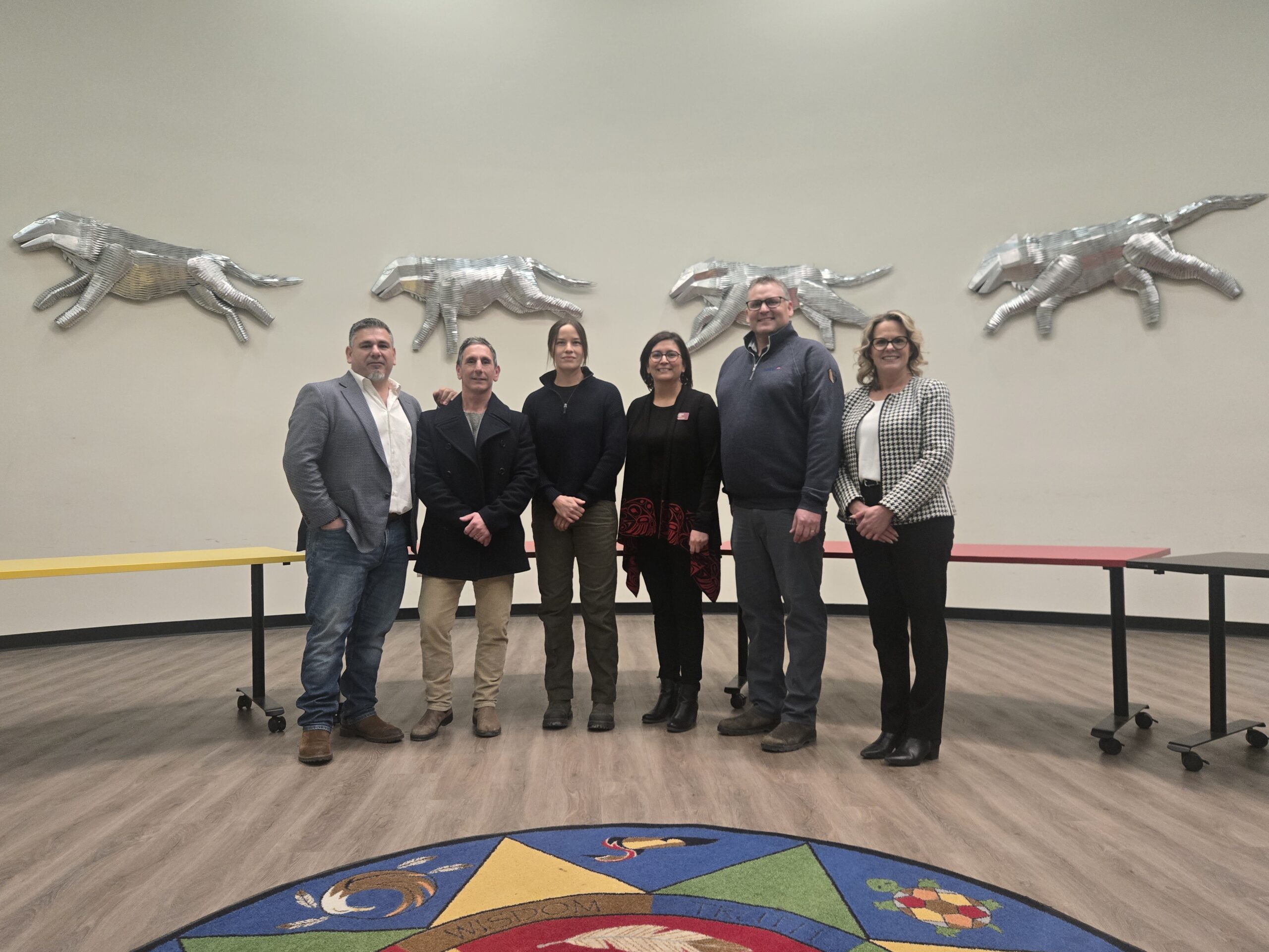 Six people stand in a line in a large room with silver animal sculptures on the wall and a colorful circular design on the floor.