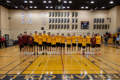 The Cambrian's men's volleyball team in matching yellow jerseys stands side by side on an indoor gym court with arms around each other.