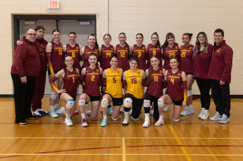 The Cambrian's women’s volleyball team poses indoors with two coaches, wearing maroon and gold Cambrian jerseys, standing and kneeling on a gym floor.