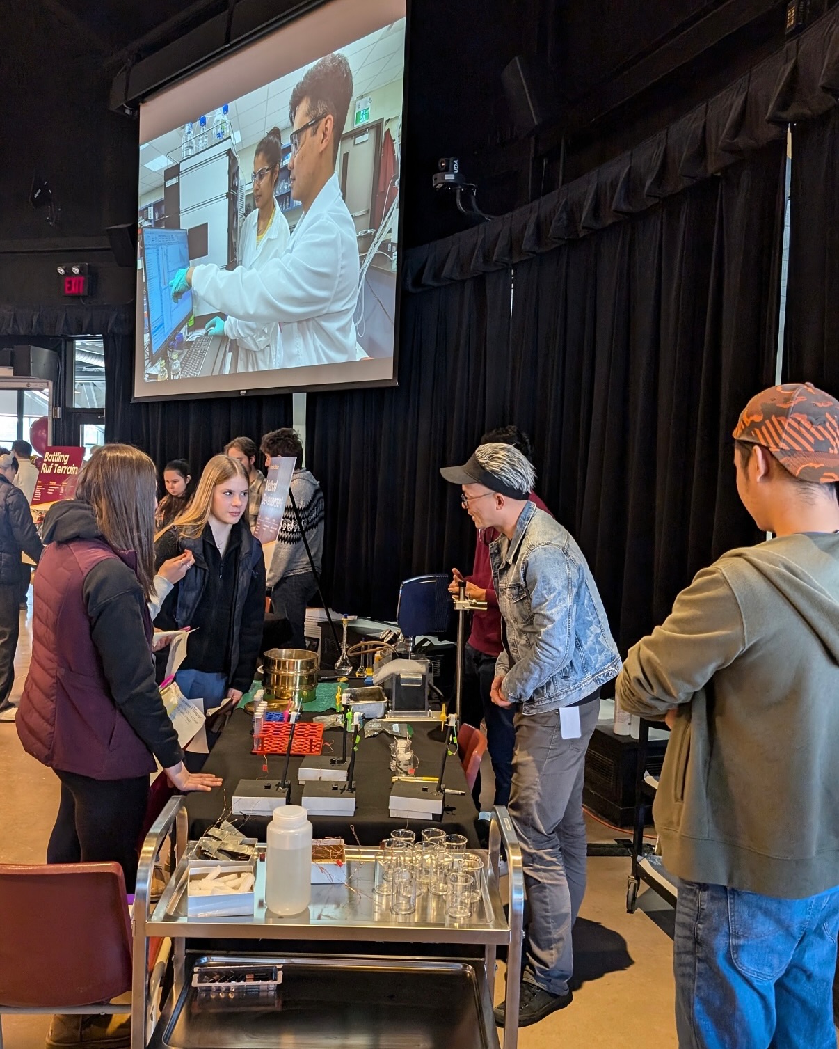 People interact at a science fair booth with equipment on a table; a screen above shows a lab scene with two scientists working together.