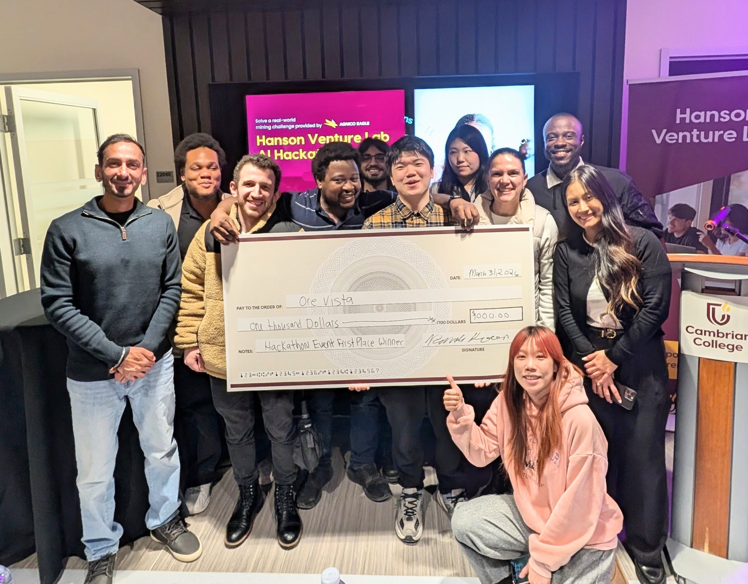 A group of people poses indoors holding a large novelty check for $1,000, awarded for a first-place win at a Hanson Venture Hub Hackathon.