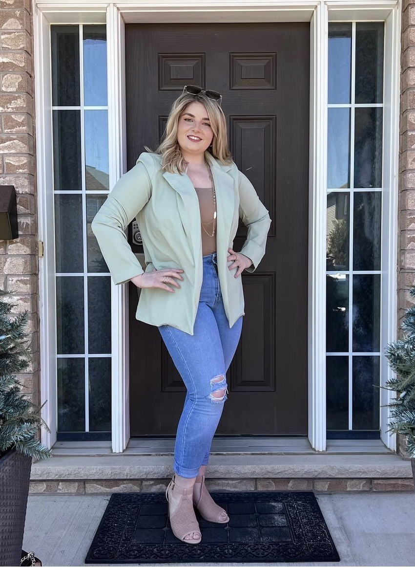 Stephanie Hutton stands in front of a brown door with glass panels, wearing a light green blazer, tan top, ripped blue jeans, and beige shoes, smiling at the camera.