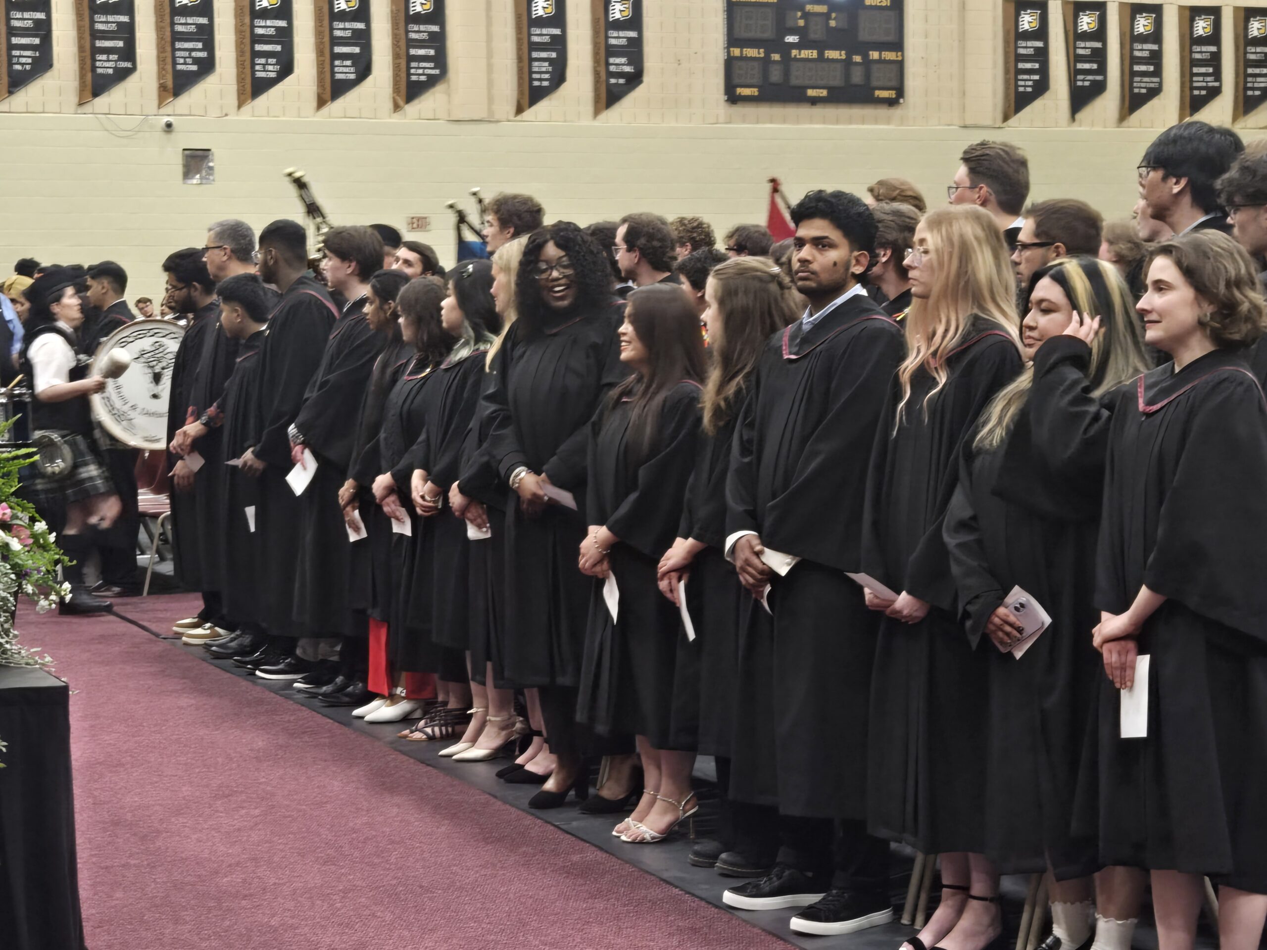 A group of graduates in black robes stand in rows during a graduation ceremony inside a gymnasium.